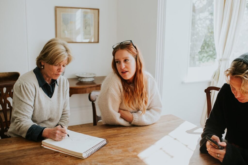 Three woman sit around a dining table in a sunny house, making notes