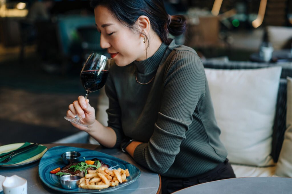 Beautiful young Asian woman drinking glass of red wine while eating beef steak in restaurant. Luxury lifestyle. People, food and lifestyle